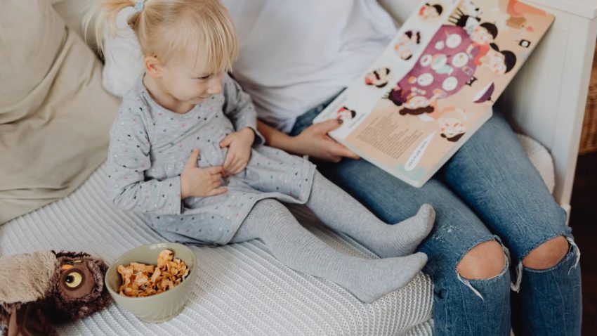 Mother reading a book to her daughter in a cozy bedroom setting with snacks nearby.